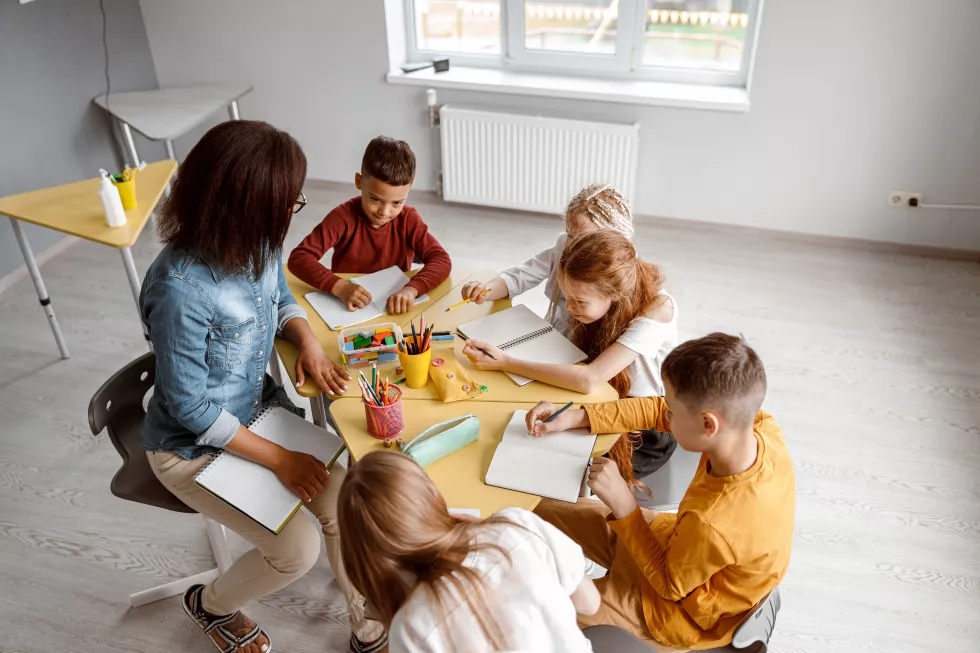 children-writing-notebooks-while-sitting-table-with-teacher