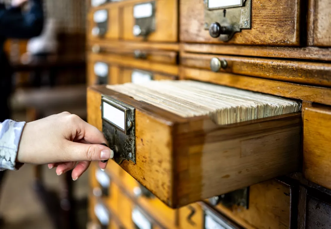 Dłoń otwiera szufladę drewnianego katalogu kartkowego w bibliotece.