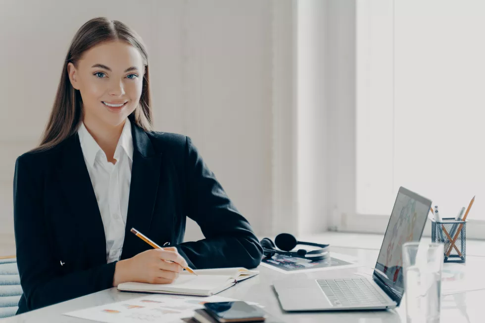 portrait-smiling-young-caucasian-bussiness-woman-black-formal-suit-looking-camera-sitting-by-big-white-desk-with-note-book-laptop-window-light-minimalistic-background