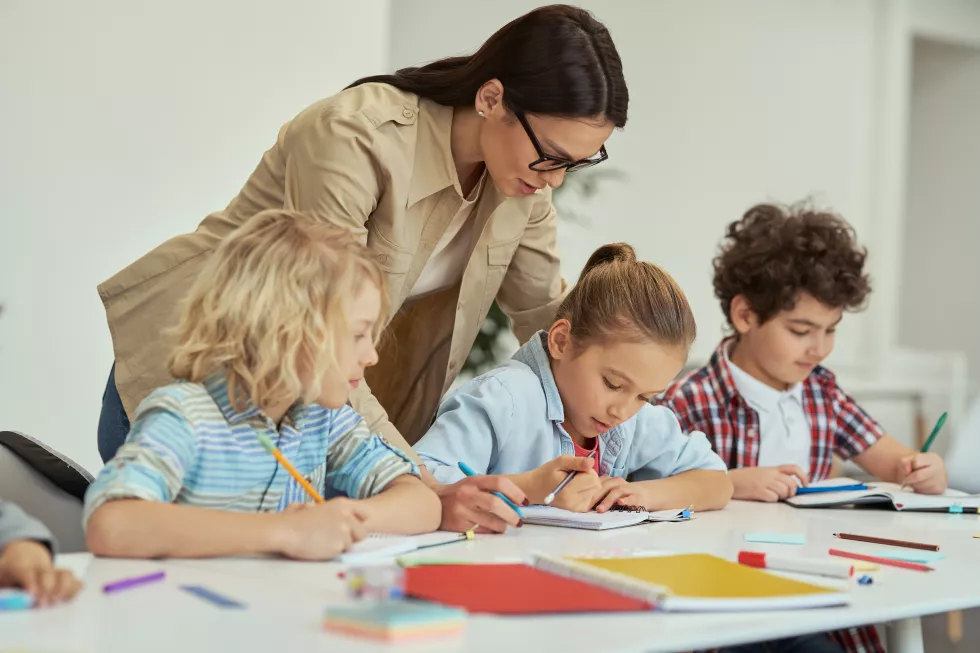 excellence-kind-young-female-teacher-glasses-helping-her-little-schoolchildren-classroom