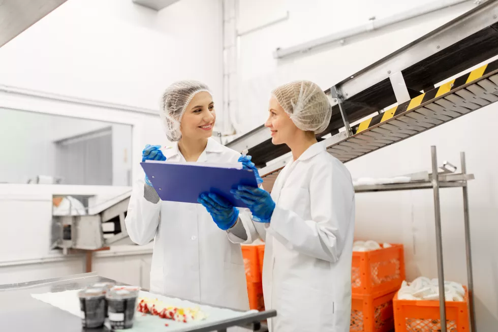 food-production-industry-people-concept-happy-women-technologists-with-clipboard-tasting-ice-cream-quality-factory