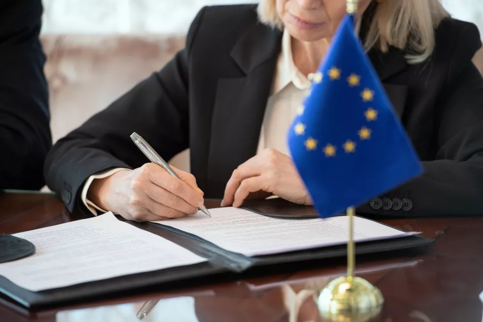flag-european-union-table-against-mature-female-delegate-signing-contract-after-negotiating-making-deal-by-table-boardroom(1)