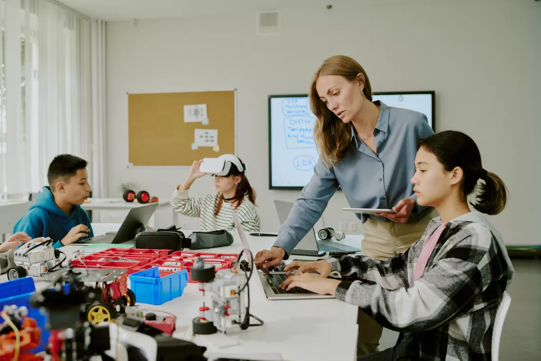 female-teacher-with-tablet-helping-young-programmer-with-codes-while-other-students-enjoying-vr