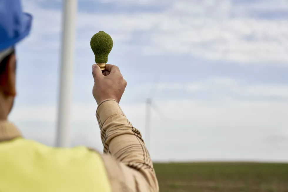 hand-unrecognizable-man-holding-green-electric-bulb-wind-turbine-background